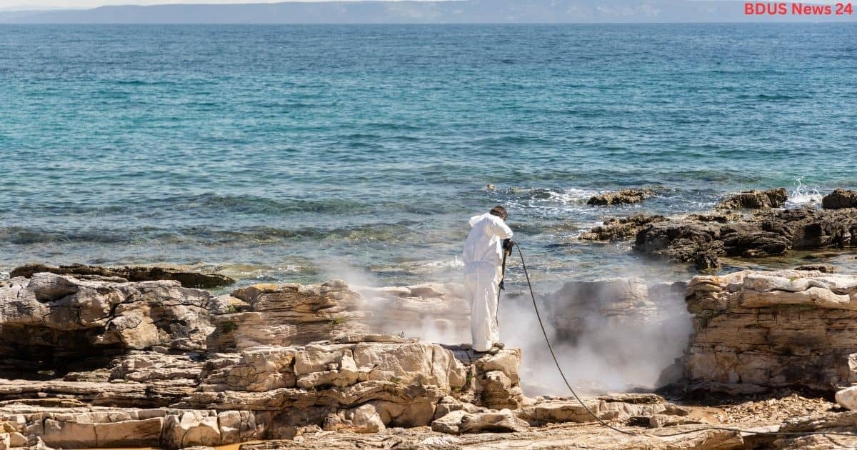 Person in white protective suit pressure washing rocks on the coast
