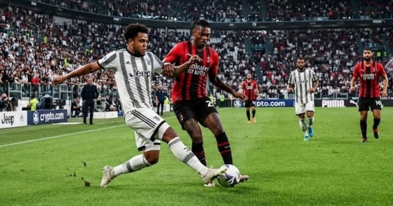 A football match action shot between Juventus and AC Milan players at a packed stadium, with a Juventus player in a white and black striped kit tackling an AC Milan player in a red and black striped kit.